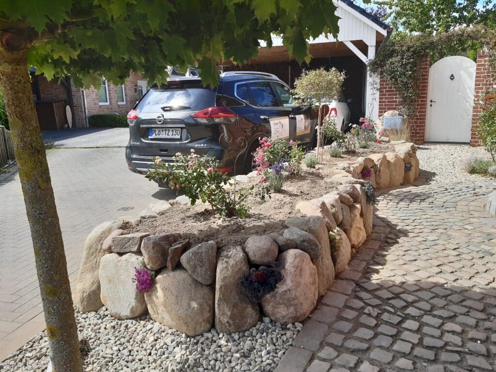 Ein kleines Steingartenbeet, gestaltet durch sorgfältige Gartengestaltung und Gartenpflege, zeigt verschiedene Blumen und Pflanzen zwischen großen Steinen. Dahinter parkt ein schwarzer Geländewagen vor einem Haus mit Backsteinmauer und weißem Garagentor.