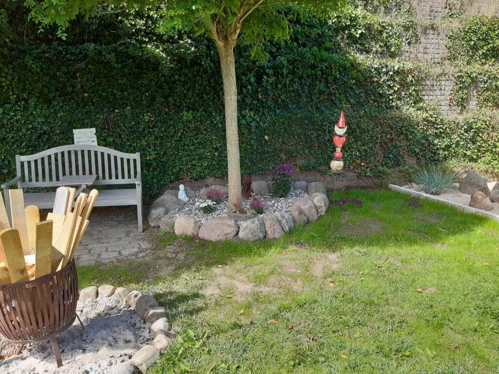 Eine Gartenszene mit einer weißen Bank, einem von Steinen und Blumen umgebenen Baum, einer rot-weißen Gnom-Statue, einer Feuerstelle mit Holzteilen und grünem Gras, das von einer blattreichen Hecke begrenzt wird - perfekte Inspiration für kreative Gartengestaltung.