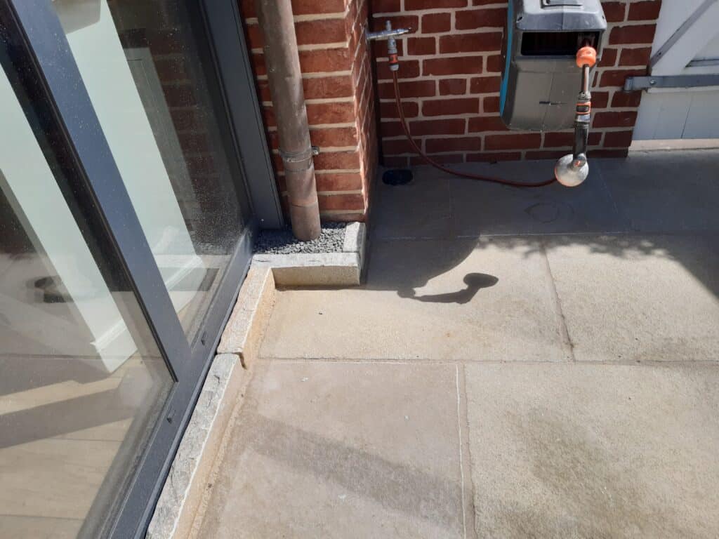 Ein sonniger Terrassenbereich mit Steinfliesen, einer Ziegelmauer, einer Glastür und einem Metallkasten ist ein Beispiel für durchdachte Gartengestaltung. Ein Abflussrohr und ein Gartenwasserhahn sorgen für zusätzlichen Nutzen, während die Schatten der Sonne diesen einladenden Raum verschönern.