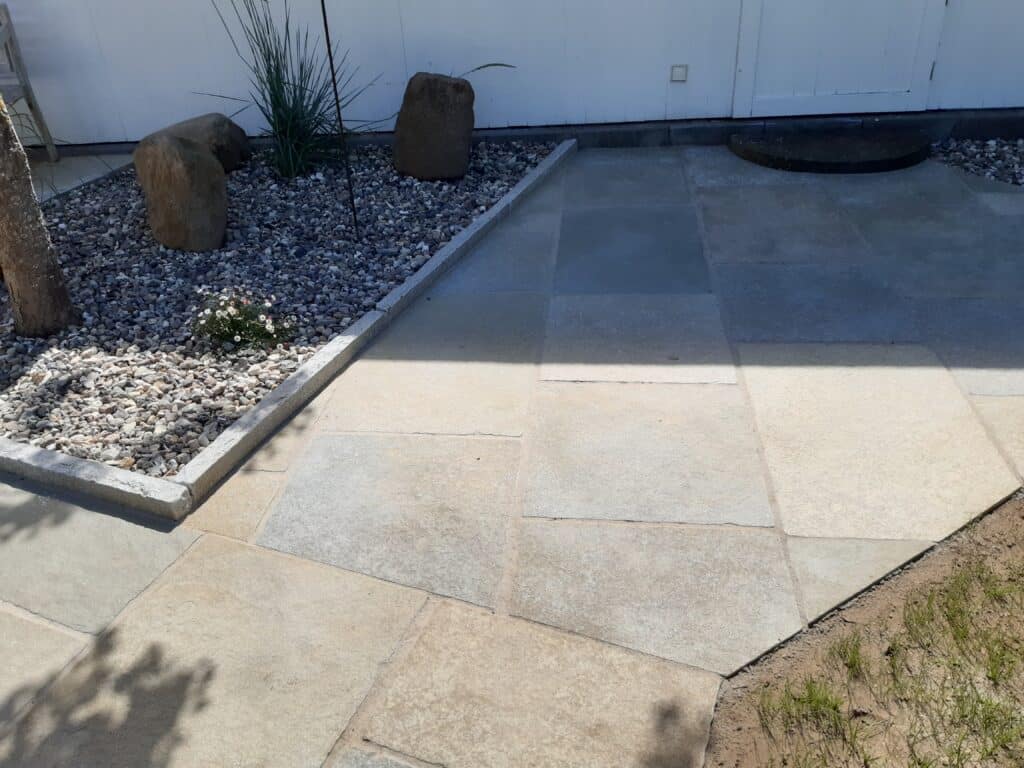 Eine gepflasterte Terrasse mit quadratischen Steinfliesen liegt neben einem kleinen Gartenbeet - eine stilvolle Gartengestaltung mit weißen Kieselsteinen, Steinen und einigen Grünpflanzen, die von einer niedrigen Betonkante begrenzt werden. Ein Teil der Fläche ist schattig.