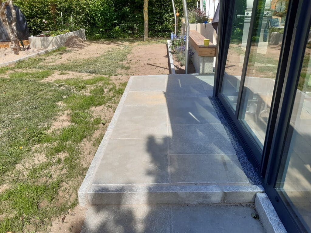 Neben einer Glastür befindet sich eine Betonterrasse mit großen Fliesen, die eine durchdachte Gartengestaltung erkennen lassen. Die Terrasse ist mit Gras und Erde eingefasst, im Hintergrund sind Pflanzen und eine kleine Stützmauer zu sehen.