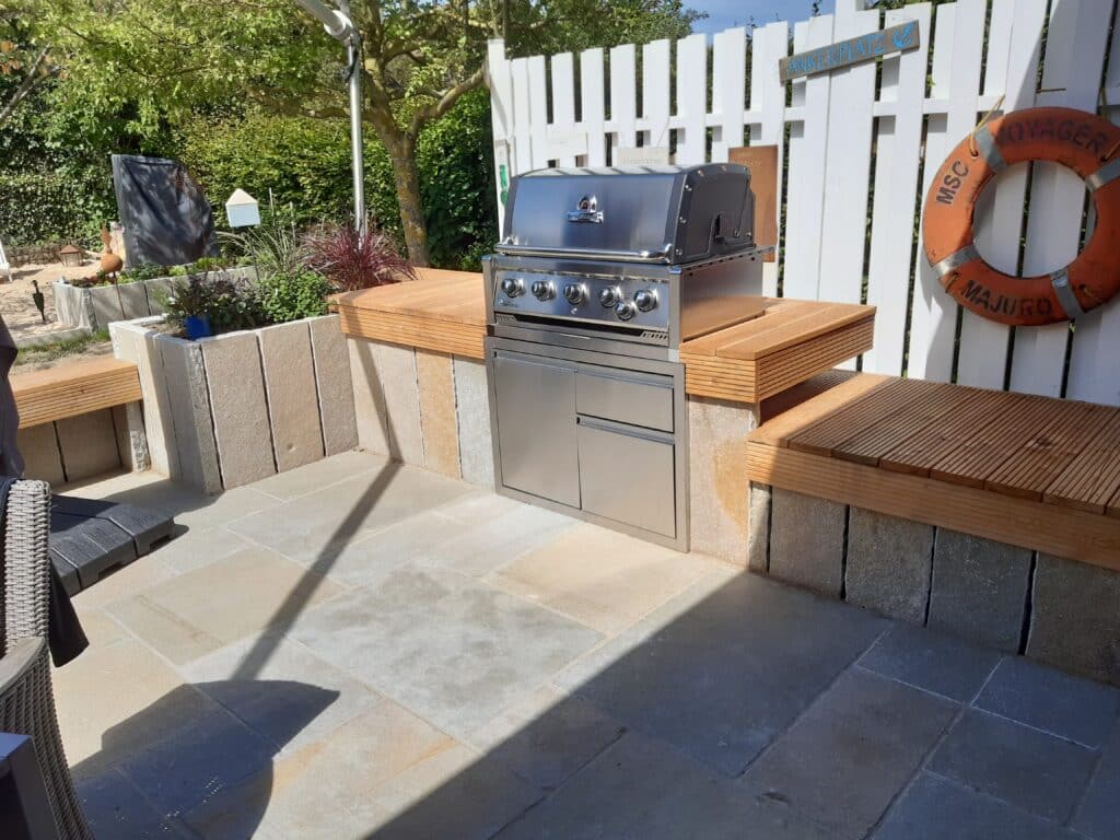 Ein Terrassenbereich mit eingebautem Gasgrill aus Edelstahl, Holzbänken, weißem Zaun, einer Rettungsboje am Zaun und üppigem Grün - perfekt, um Gartengestaltung und Gartenarbeit an einem sonnigen Tag zu genießen.
