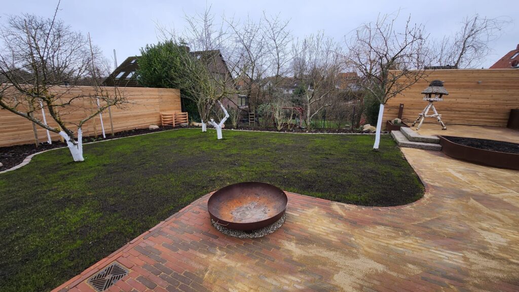 In dem von Experten gestalteten Garten befindet sich eine runde Feuerstelle auf einer gemauerten Terrasse, umgeben von einer Rasenfläche und Bäumen mit weiß gestrichenen Stämmen. Holzzäune und eine kleine Struktur verstärken die ruhige Umgebung.