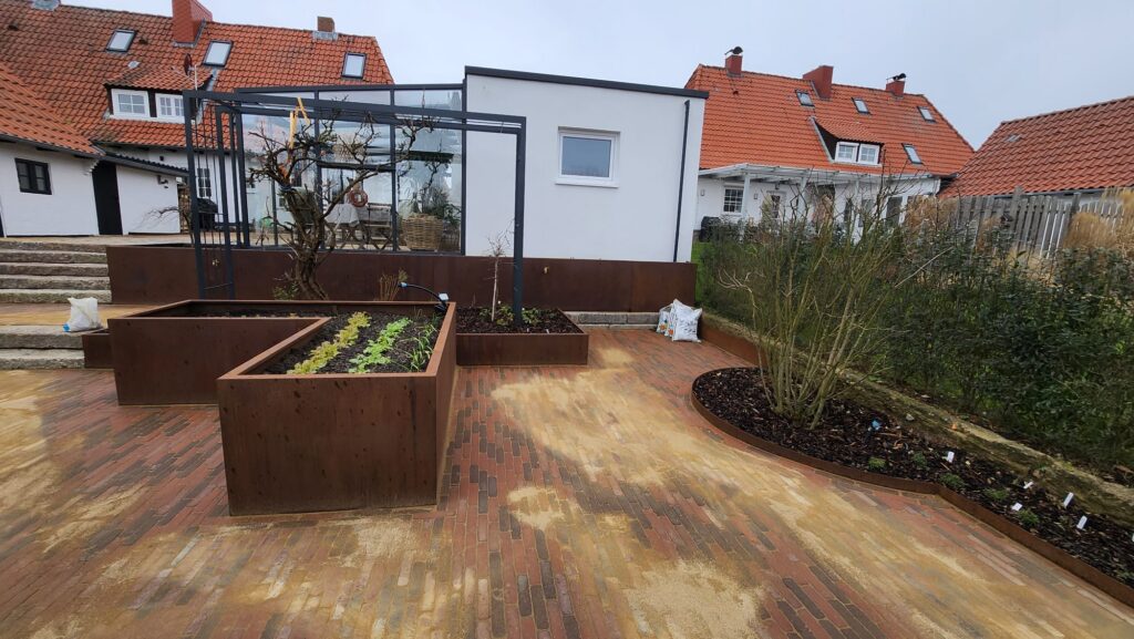Ein aufgeräumter Garten zeigt eine durchdachte Gartengestaltung mit Hochbeeten mit Jungpflanzen, einem gläsernen Gewächshaus, einem weißen Schuppen und gemauerten Wegen vor dem Hintergrund roter Ziegeldachhäuser unter einem bedeckten Himmel.