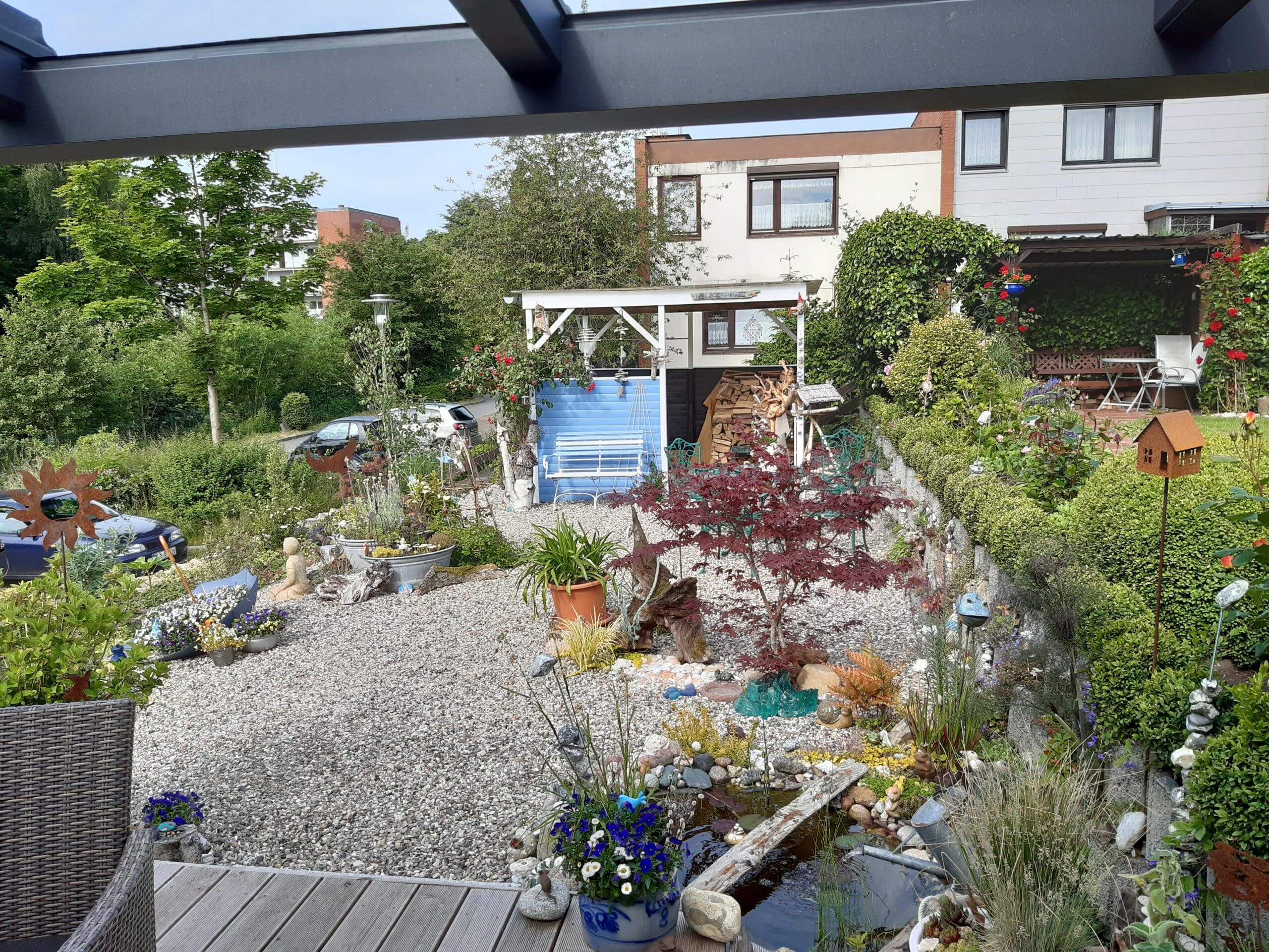 Ein gepflegter Garten mit Kieswegen, farbenfrohen Blumen, einem rotblättrigen Baum, einem kleinen Teich, Vogelhäuschen und geschmackvollen Dekorationen dient als inspirierende Referenz für die Gestaltung des Außenbereichs. Eine weiße Bank unter einer Pergola und Terrassenmöbel vervollständigen das einladende Bild.