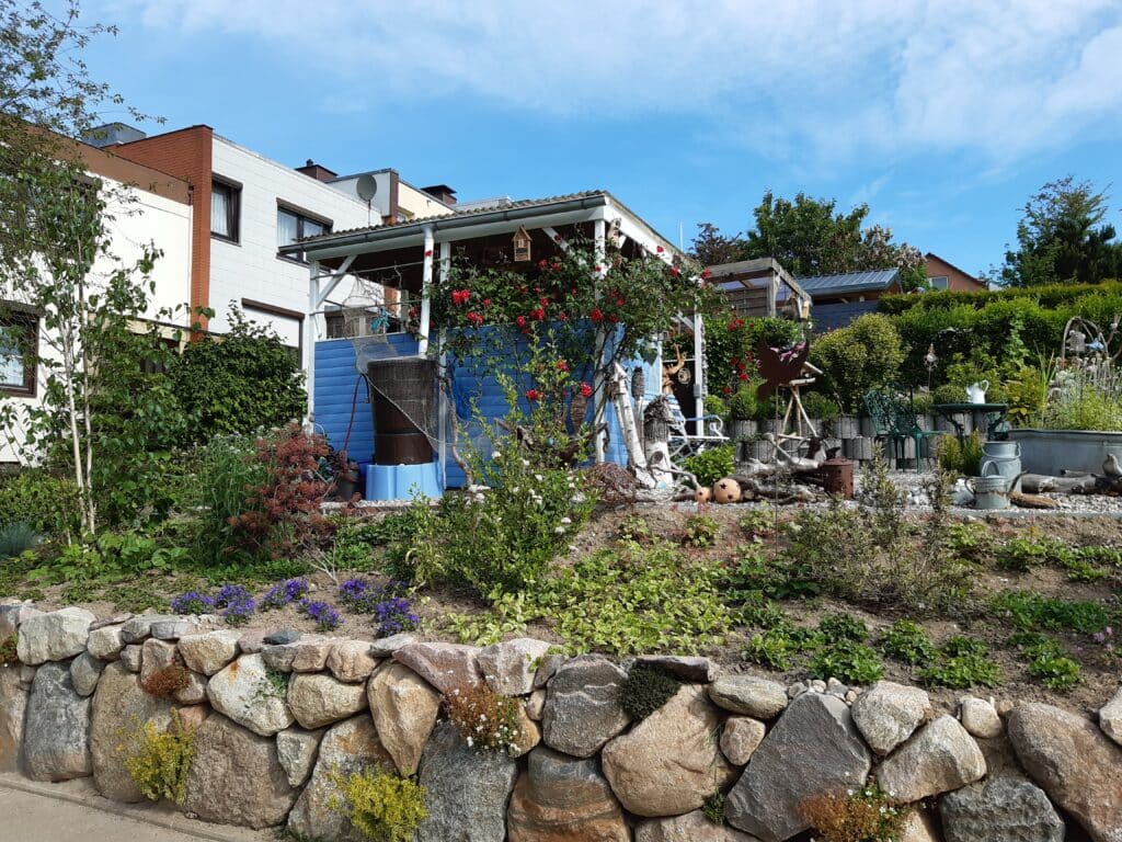 Ein bunter Garten mit verschiedenen Pflanzen und Blumen vor einem kleinen blauen Gartenhäuschen dient als eines unserer charmanten Referenzen. Die Szene wird von einer Steinmauer begrenzt, und unter dem blauen Himmel sind weiße und gemauerte Häuser zu sehen.