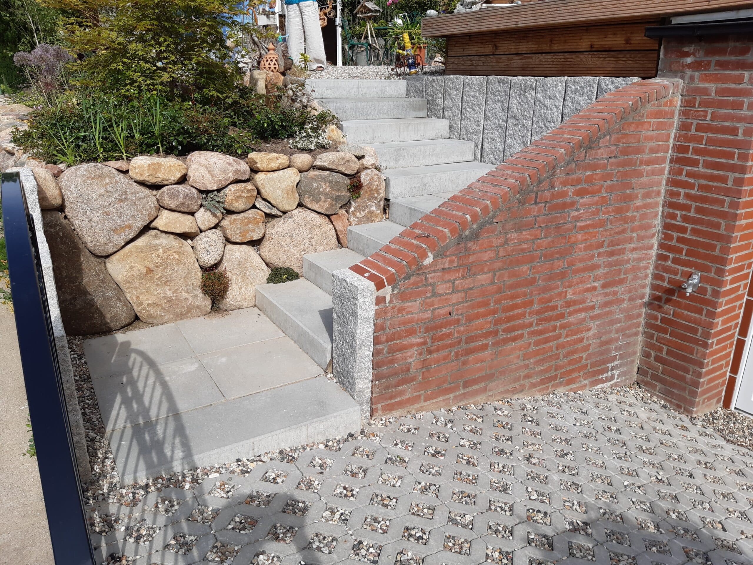 Eine von einer roten Backsteinmauer eingefasste Betontreppe führt zu einem Gartenbereich mit großen Steinen und Pflanzen. Der Boden ist mit Pflastersteinen und Schotter bedeckt.