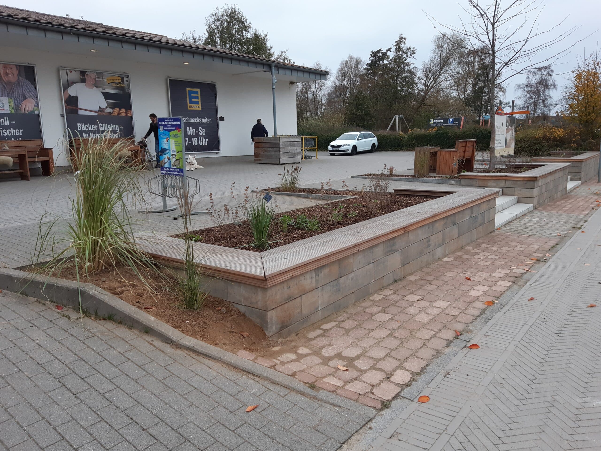 Rechteckige Hochbeete mit Holzbänken säumen eine gepflasterte Fläche vor einem Gebäude und dienen als praktische Referenzen für die Landschaftsgestaltung. Im Hintergrund sind ein weißes Auto und mehrere Personen in der Nähe eines Geschäftseingangs zu sehen.