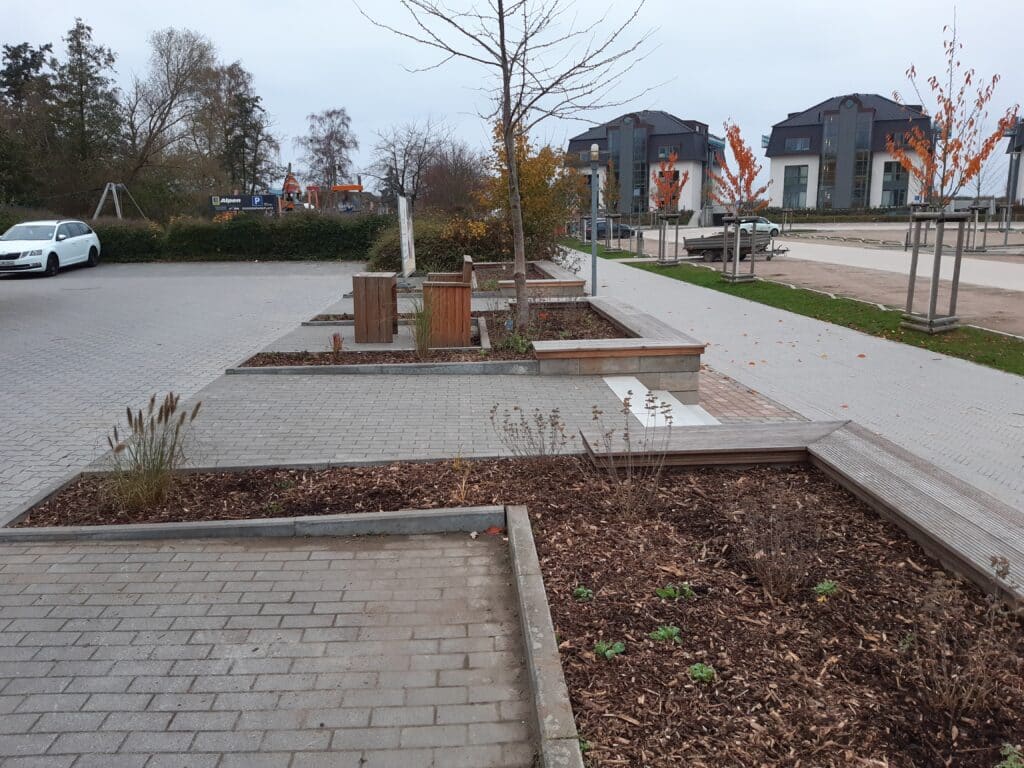 Ein moderner, gepflasterter Parkplatz mit erhöhten hölzernen Pflanzgefäßen, Bänken und jungen Bäumen an einem bewölkten Tag; geparkte Autos und moderne Wohngebäude in der Nähe dienen als Referenzen für die Gestaltung der Stadtlandschaft.