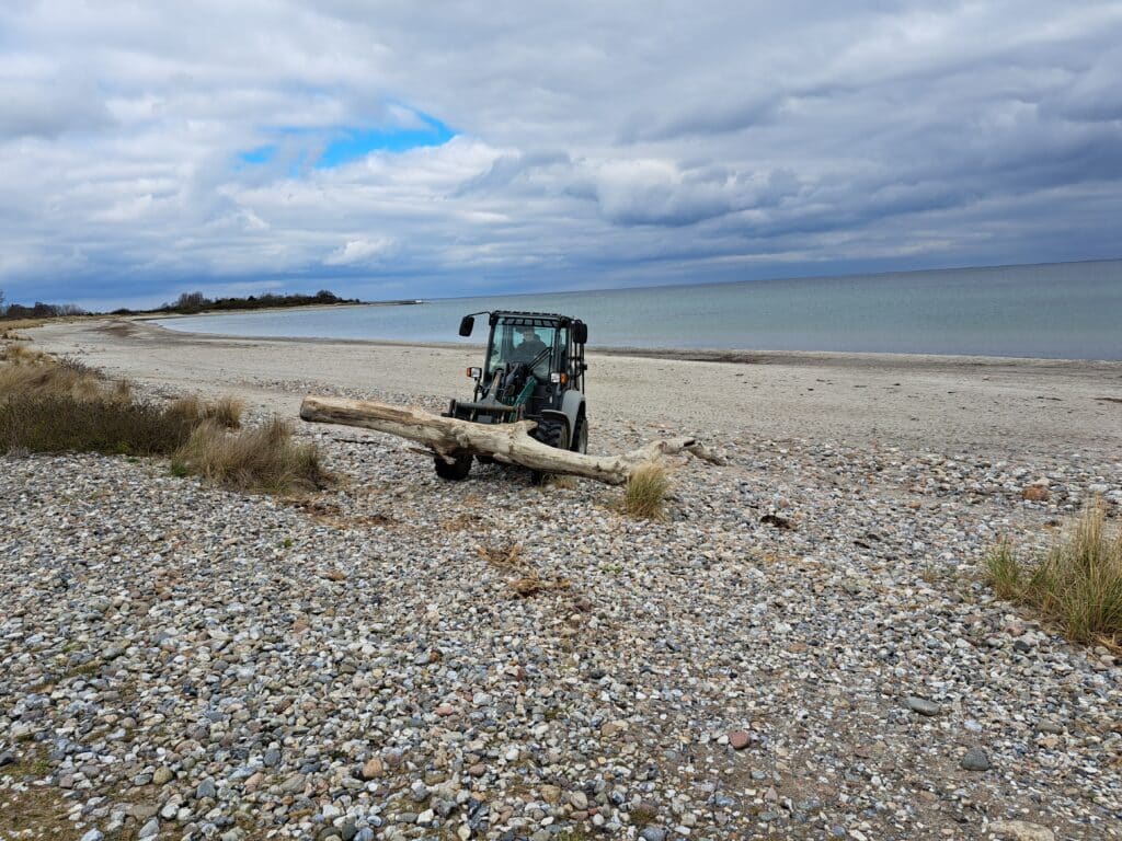Ein kleines Fahrzeug hebt einen großen Baumstamm auf einen felsigen Strand unter einem wolkenverhangenen Himmel, mit dem Meer und der fernen Küstenlinie im Hintergrund - eine beeindruckende Szene für Ihre Referenzen-Sammlung.