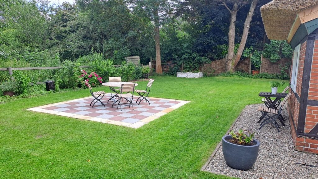 Ein Garten im Hinterhof mit grünem Rasen, einer Terrasse mit einem karierten Fliesenboden und einem runden Tisch mit vier Stühlen. Zu den Referenzen gehören ein kleiner Beistelltisch und zwei Stühle in der Nähe des Backsteinhauses, mit Bäumen und Sträuchern im Hintergrund.