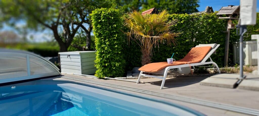 Ein Liegestuhl mit einem orangefarbenen Kissen und einem Getränk steht an einem sonnigen Tag neben einem Swimmingpool, umgeben von grünen Büschen und einer kleinen Palme - eine einladende Umgebung, die Design-Referenzen würdig ist.