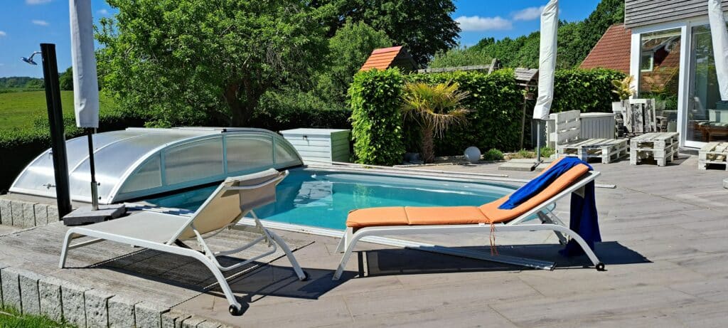 Ein teilweise überdachter Pool im Garten ist von einer Holzterrasse umgeben. Zwei Loungesessel, einer davon mit einem orangefarbenen Kissen und einem blauen Handtuch drapiert, sitzen neben dem Pool - ein einladender Ort, der stilvolle Referenzen für das Leben im Freien präsentiert. Üppiges Grün umrahmt das moderne Haus.