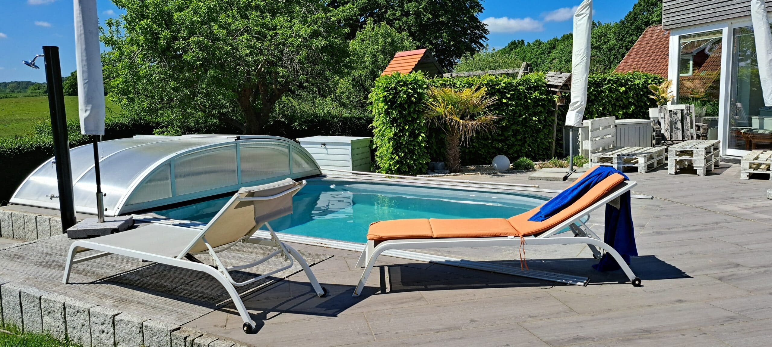 Ein teilweise überdachter Pool im Garten ist von einer Holzterrasse umgeben. Zwei Loungesessel, einer davon mit einem orangefarbenen Kissen und einem blauen Handtuch drapiert, sitzen neben dem Pool - ein einladender Ort, der stilvolle Referenzen für das Leben im Freien präsentiert. Üppiges Grün umrahmt das moderne Haus.