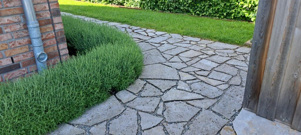 Gekrümmter Steinweg, eingefasst von grünen Sträuchern und Gras, neben einer Backsteinmauer mit einem Metallabflussrohr und einer Holzkonstruktion auf der rechten Seite. Üppiges Grün im Hintergrund, das schöne Gartenreferenzen zeigt.