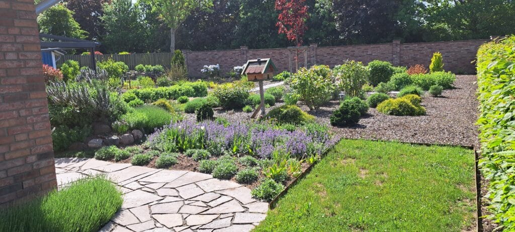 Ein ordentlich angelegter Garten mit Steinwegen, saftig grünem Gras, verschiedenen Sträuchern und blühenden Pflanzen. In der Mitte steht ein hölzernes Vogelfutterhaus, das von Mulch und einer Ziegelmauer eingefasst ist - eine inspirierende Kulisse für landschaftliche Referenzen.