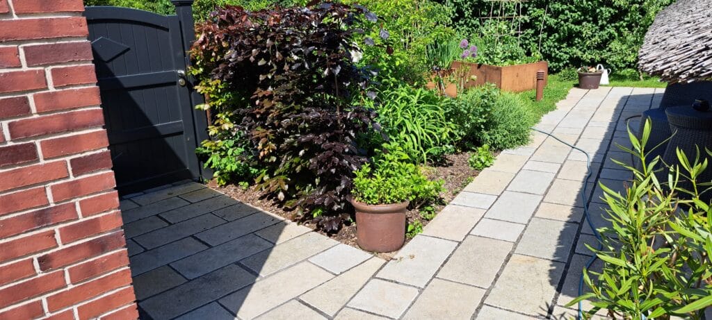 Ein sonnenbeschienener Garten mit einem gepflasterten Steinweg, Blattpflanzen, einem Topfstrauch, Hochbeeten aus Holz und einem schwarzen Tor an einer roten Backsteinmauer - ein einladender Außenbereich, der als inspirierende Referenzen für jede Landschaftsgestaltung dienen könnte.