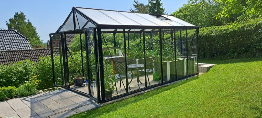 Ein gläsernes Gewächshaus steht auf einer gefliesten Terrasse in einem üppigen Garten, gefüllt mit Topfpflanzen und einem kleinen schwarzen Metalltisch mit zwei Stühlen. Umgeben von Bäumen und Hecken unter einem sonnigen Himmel, bietet es inspirierende Referenzen für jeden Gartenliebhaber.
