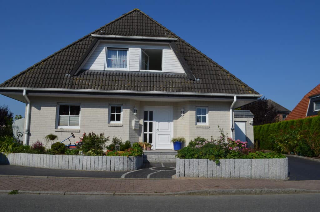 Ein weißes, zweistöckiges Haus mit einem dunklen, steil abfallenden Dach, einer Dachgaube und einem gepflegten, von niedrigen Steinmauern eingefassten Vorgarten vor einem strahlend blauen Himmel - ein beeindruckendes Beispiel unter unseren Referenzen.