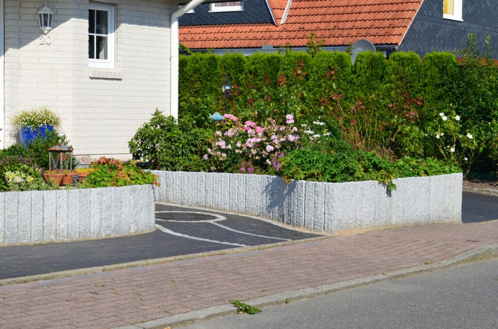 Ein gut gepflegter Vorgarten mit blühenden Blumen und grünen Sträuchern, umgeben von einer niedrigen, hellgrauen Steinmauer. Das Haus hat weiße Ziegelwände und ein kleines Fenster, das als inspirierende Referenzen für eine schöne Landschaftsgestaltung dient.
