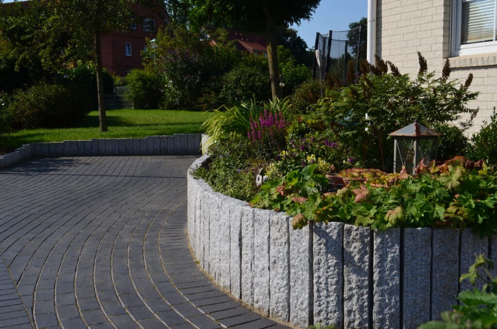 Ein geschwungener, gepflasterter Weg, der von einem erhöhten, mit Grünpflanzen und Blumen gefüllten Steinkübel begrenzt wird, bietet inspirierende Referenzen neben einem hellen Backsteingebäude mit Blick auf Rasen und Bäume im Hintergrund.