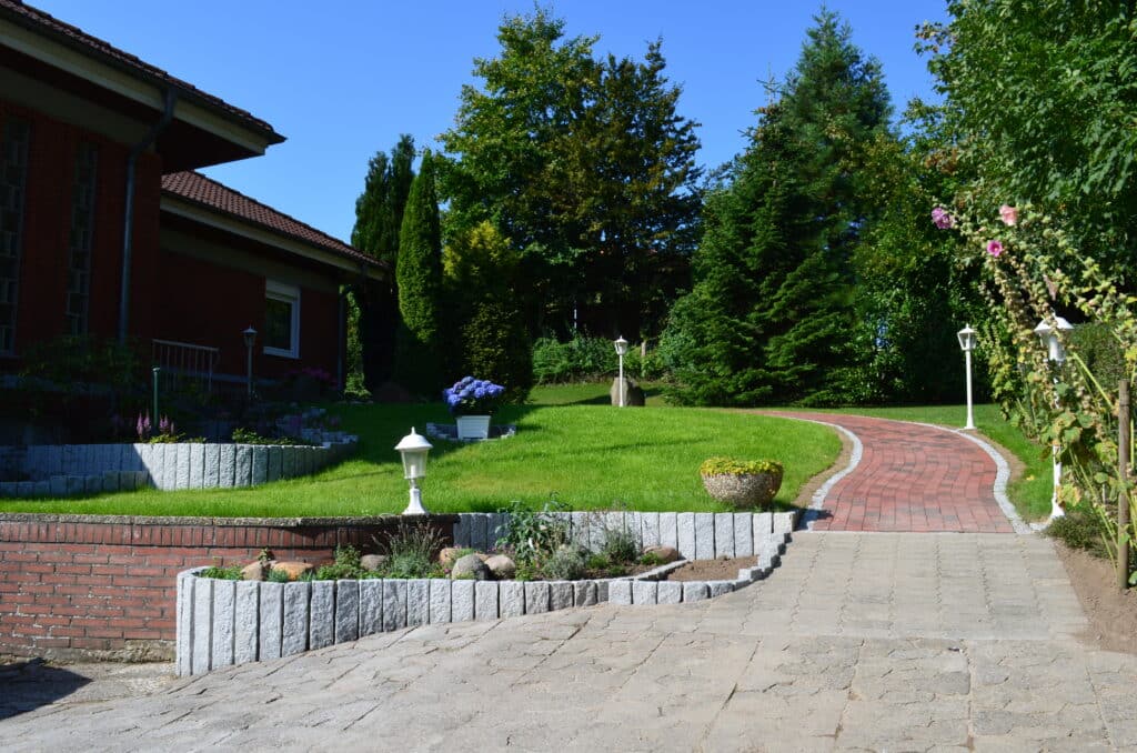 Ein gemauerter Weg schlängelt sich durch einen ordentlich angelegten Garten mit grünem Gras, Bäumen, Sträuchern und weißen Laternenpfählen - eine reizvolle Szene, die man in unseren Referenzen oft sieht. Auf der linken Seite ist ein rotes Backsteinhaus unter einem klaren blauen Himmel zu erkennen.