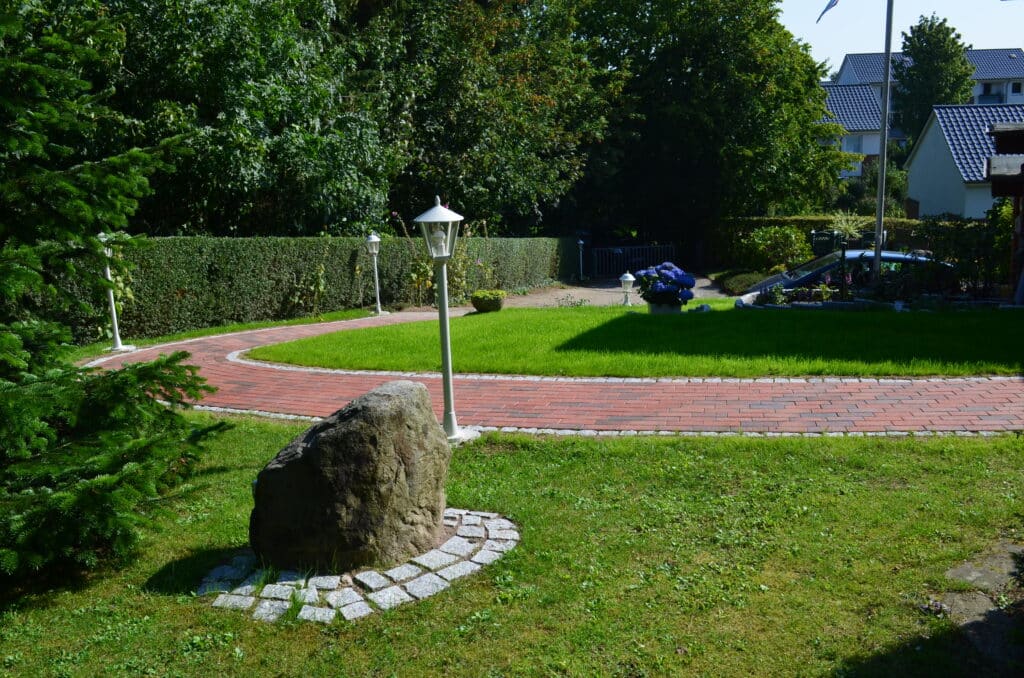 Ein gepflegter Garten mit einem geschwungenen Ziegelweg, Rasen, Hecken, einem großen Felsen, weißen Laternenpfählen und Bäumen ist ein schönes Beispiel für die Landschaftsgestaltung in der Nähe von Wohnhäusern unter freiem Himmel.