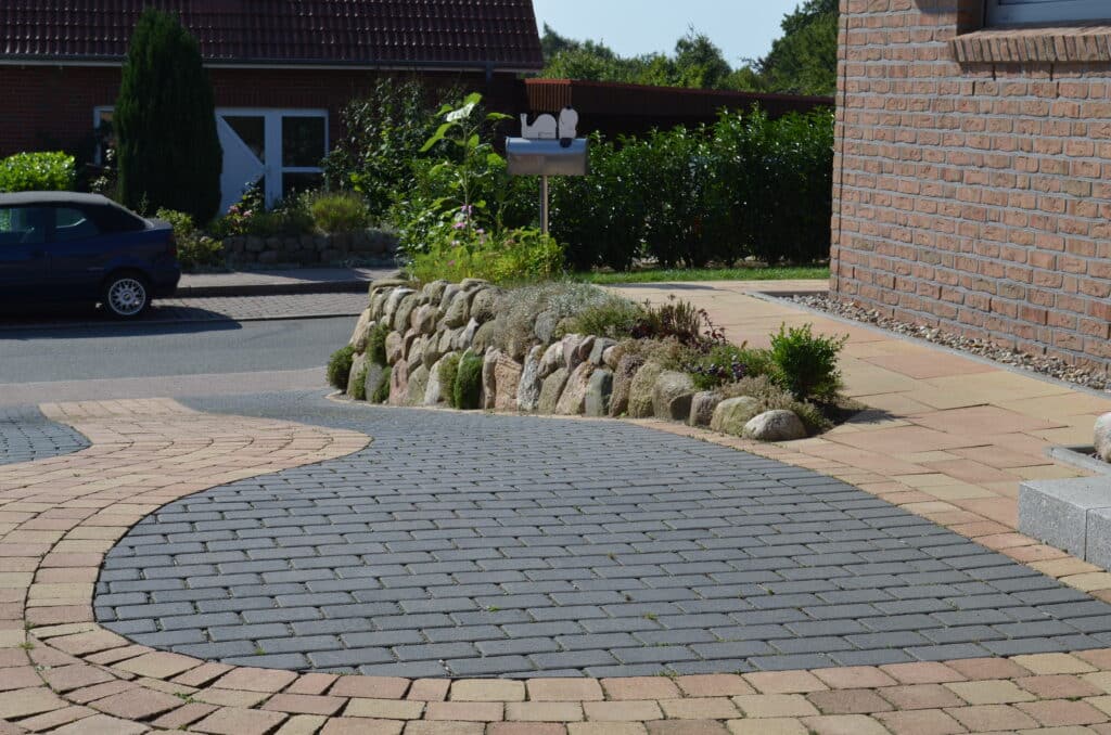Gebogene Einfahrt mit grauen und hellbraunen Pflastersteinen, eingefasst von einer niedrigen Steinmauer mit Pflanzen und einem gepflegten Garten. Rechts ein Backsteinhaus, ein auf der Straße geparktes Auto, im Hintergrund ein weiteres Haus und grüne Hecken.