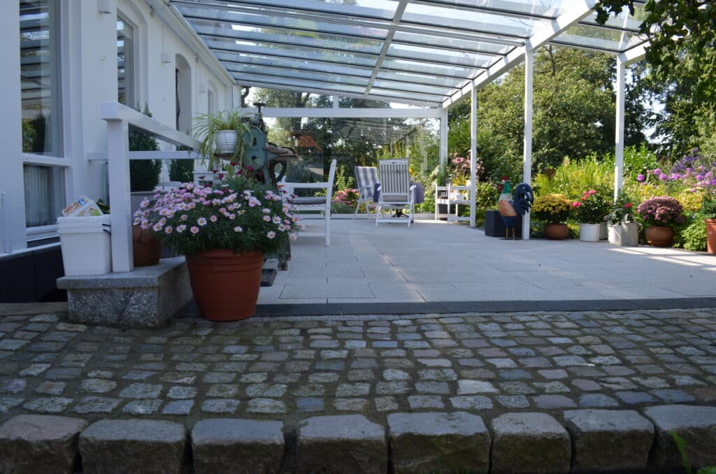 Eine sonnenbeschienene Terrasse mit einem Glasdach, weißen Stühlen und Topfblumen am Rande. Ein gepflasterter Weg führt zum gefliesten Sitzbereich, der von Grünpflanzen und farbenfrohen Referenzen und Dekorationen für den Außenbereich umgeben ist.