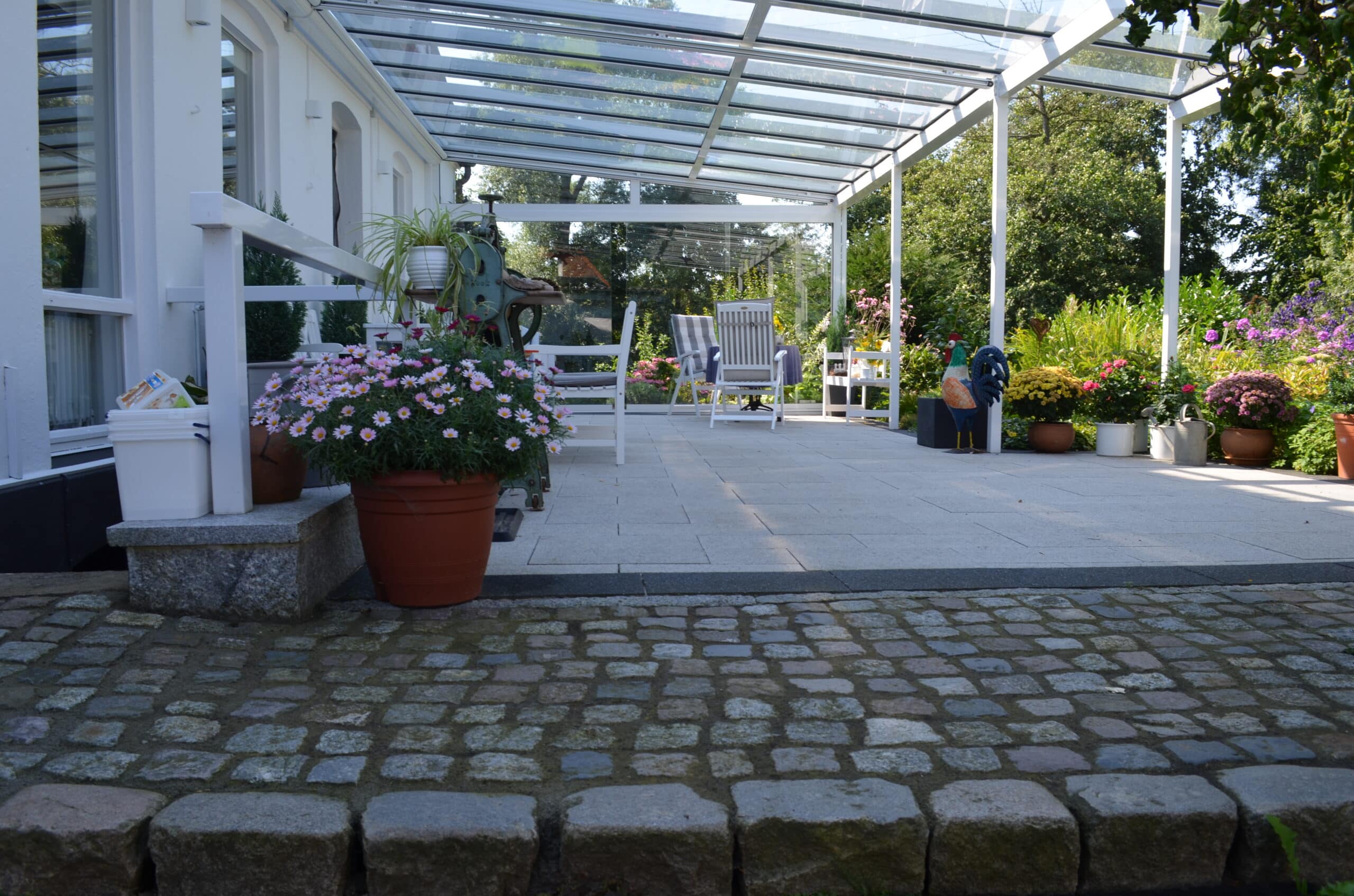Eine sonnenbeschienene Terrasse mit einem Glasdach, weißen Stühlen und Topfblumen am Rande. Ein gepflasterter Weg führt zum gefliesten Sitzbereich, der von Grünpflanzen und farbenfrohen Referenzen und Dekorationen für den Außenbereich umgeben ist.