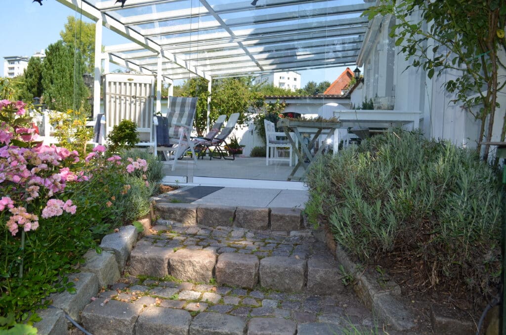 Steinstufen führen zu einer Terrasse mit Gartenmöbeln unter einem Glasdach. Rosa Blumen und Blattpflanzen säumen die Stufen, während weiße Gebäude eine elegante Kulisse bilden - eines unserer Lieblingsreferenzen für stilvolles Wohnen im Freien.