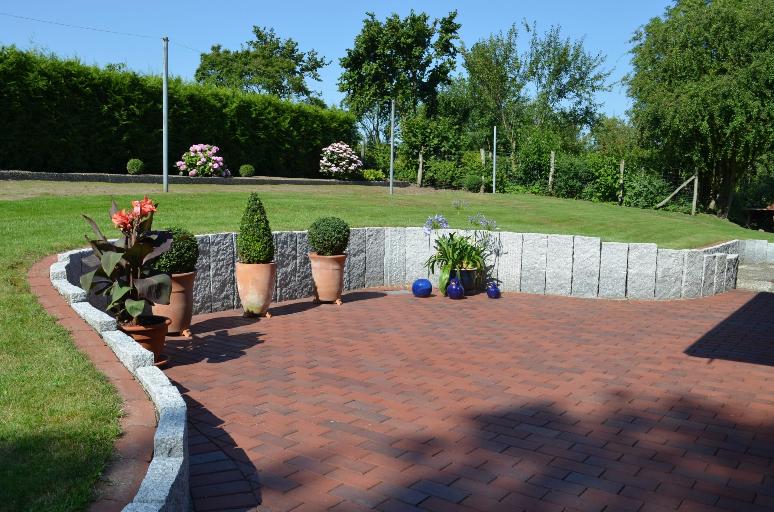 Eine sonnige, mit roten Ziegeln gepflasterte Terrasse, die von einer geschwungenen Steinkante begrenzt wird, beherbergt mehrere Topfpflanzen und blaue Vasen entlang des Randes - eine perfekte Szene für Designreferenzen. Grüner Rasen und Bäume rahmen den Hintergrund unter einem klaren blauen Himmel ein.