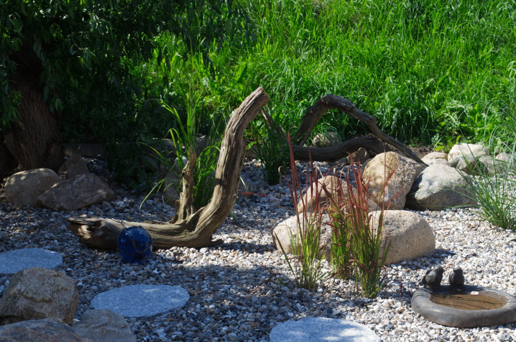 Ein kleiner Garten mit großen Steinen, Kies, trockenem Treibholz, Grasbüscheln und einer Vogeltränke, aus der zwei schwarze Vögel trinken, bietet subtile Referenzen an traditionelle Naturlandschaften vor einem Hintergrund aus dichtem grünen Laub.