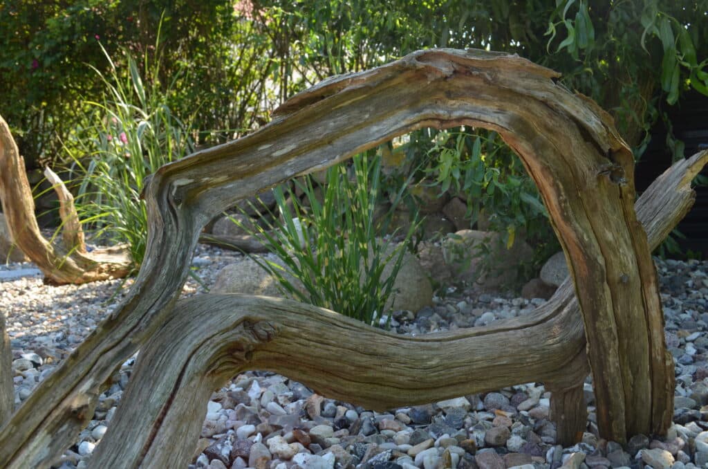 Ein verbogenes, verwittertes Stück Treibholz liegt auf einem Bett aus kleinen Steinen und Kieseln, mit grünen Pflanzen und Büschen im Hintergrund an einem sonnigen Tag - perfekt für von der Natur inspirierte Referenzen.
