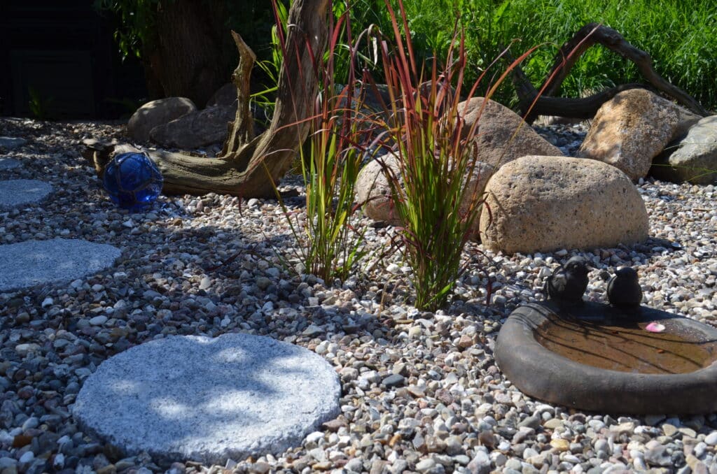 Sonnenbeschienener Garten mit runden Trittsteinen auf Kies, hohem Gras, Felsen, einer blauen Zierkugel, Treibholz und einem kleinen Vogelbad mit zwei Vogelfiguren. Grünes Laub im Hintergrund inspiriert zu Ihren Referenzen.