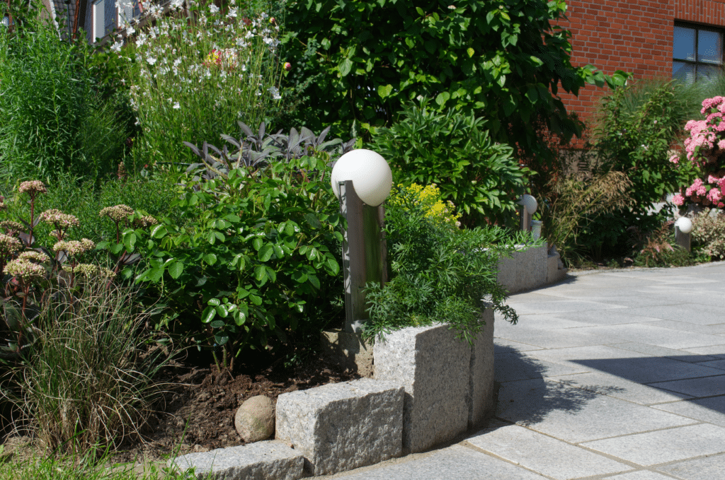 Ein Garten mit üppigen grünen Pflanzen und Blumen, eingefasst von einer Steineinfassung; moderne runde Gartenleuchten säumen den gepflasterten Weg neben einem roten Backsteingebäude und zeigen Designreferenzen für stilvolle Außenbereiche.