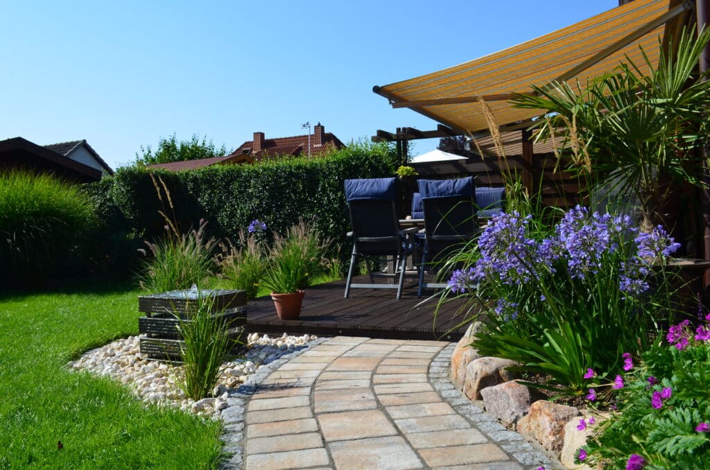 Ein geschwungener Steinweg führt zu einer Holzterrasse mit zwei Stühlen und einem Tisch unter einer gelb gestreiften Markise, die von grünem Gras, blühenden Pflanzen und einem kleinen Wasserspiel umgeben ist - perfekt, um an einem sonnigen Tag Ihre eigenen Referenzen zu inspirieren.