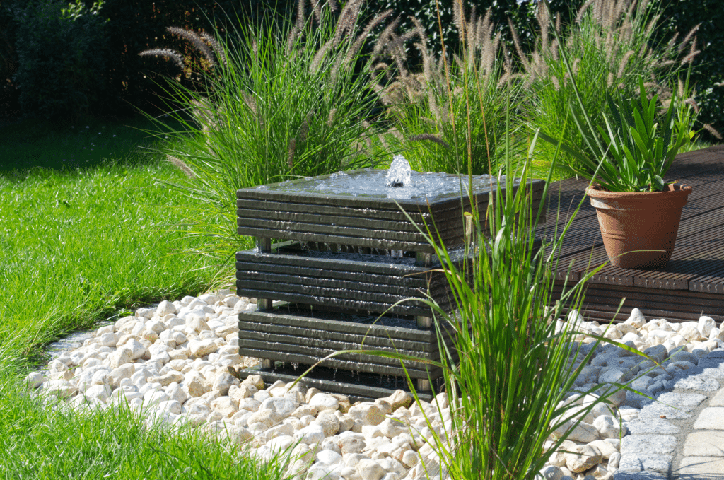 Ein moderner quadratischer Gartenbrunnen fließt über gestapelte schwarze Platten, umgeben von weißen Dekofelsen und hohem grünen Gras, und bietet stilvolle Referenzen für die Gestaltung des Außenbereichs, mit einer Topfpflanze auf einem Holzdeck im Hintergrund.