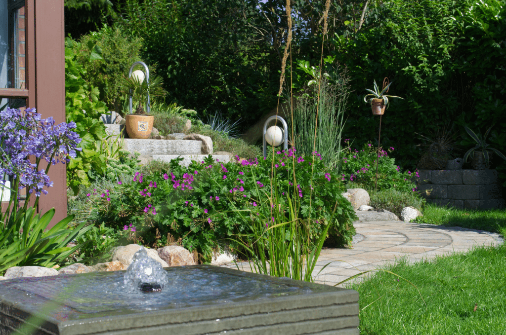 Ein sonnenbeschienener Garten mit einem sprudelnden, quadratischen Wasserbrunnen im Vordergrund, violetten Blumen und Grünpflanzen, Steinstufen, Topfpflanzen und einem geschwungenen, von üppiger Vegetation umgebenen Steinweg - eine inspirierende Kulisse für Ihre Referenzen.