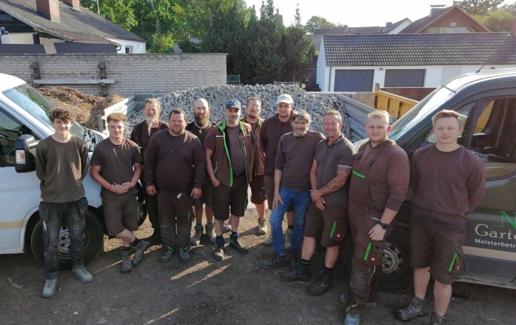 Eine Gruppe von zwölf Personen in braunen Arbeitsuniformen steht im Freien zwischen zwei Arbeitsfahrzeugen auf einer Garten- und Landschaftsbau-Baustelle, im Hintergrund sind Schotter und Häuser zu sehen.
