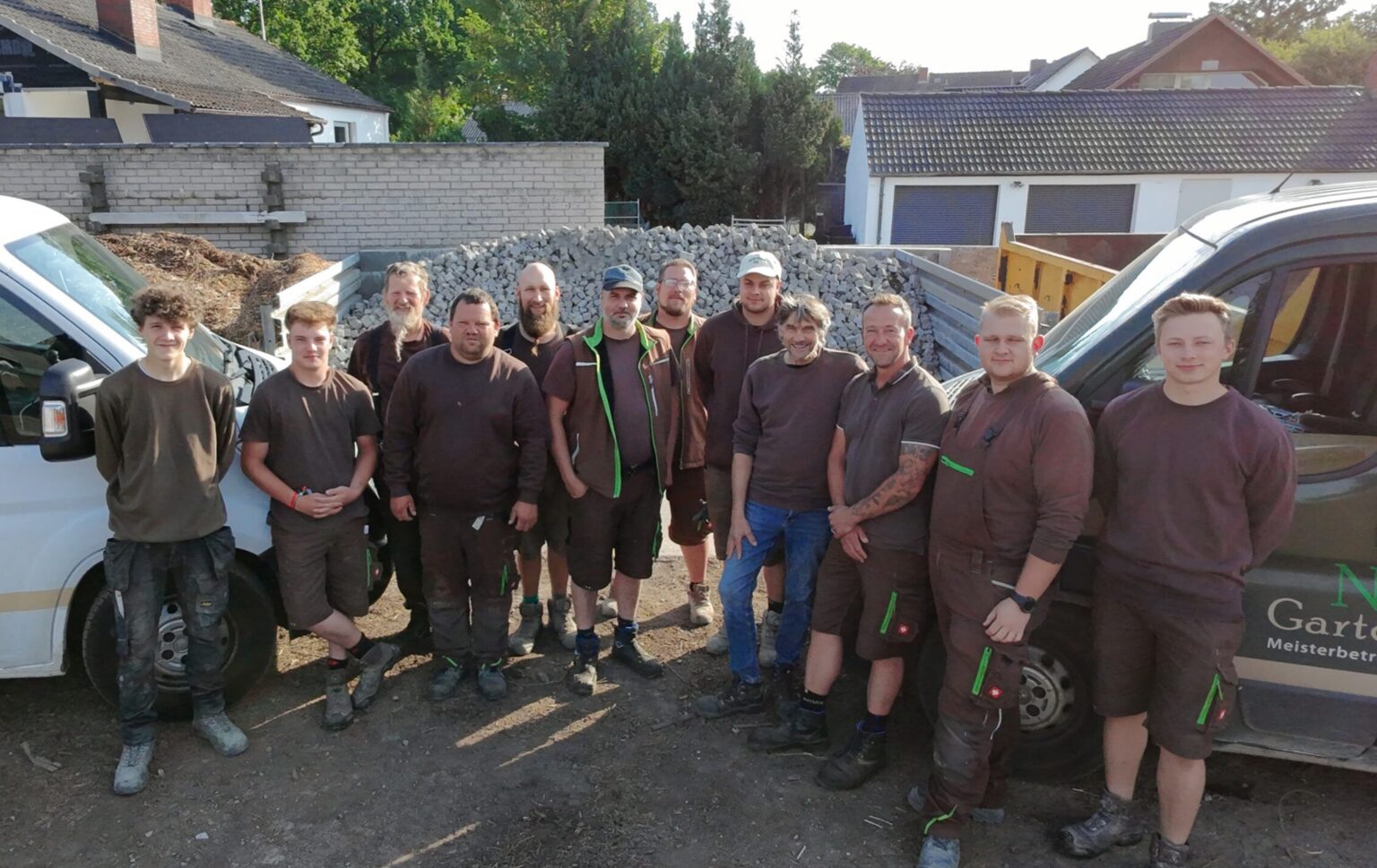 Eine Gruppe von zwölf Personen in braunen Arbeitsuniformen steht im Freien zwischen zwei Arbeitsfahrzeugen auf einer Garten- und Landschaftsbau-Baustelle, im Hintergrund sind Schotter und Häuser zu sehen.