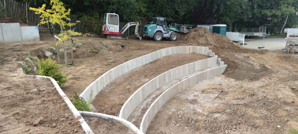 Eine Baustelle im Freien zeigt zwei geschwungene Betonstützmauern an einem unbefestigten Hang, mit Baufahrzeugen und Baumaterialien im Hintergrund, die beeindruckende Referenzen hervorheben. Ein junger Baum und einige kleine Pflanzen erscheinen auf der linken Seite des Bildes.