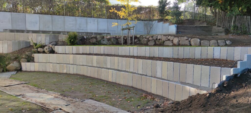 Geschwungene, abgestufte Steinstützmauern in einem mit Pflanzen, Felsen und einem jungen Baum bepflanzten Außenbereich. In unseren Referenzen finden Sie ähnliche Designs mit Hängen, Holzzäunen und üppigem Grün im Hintergrund.