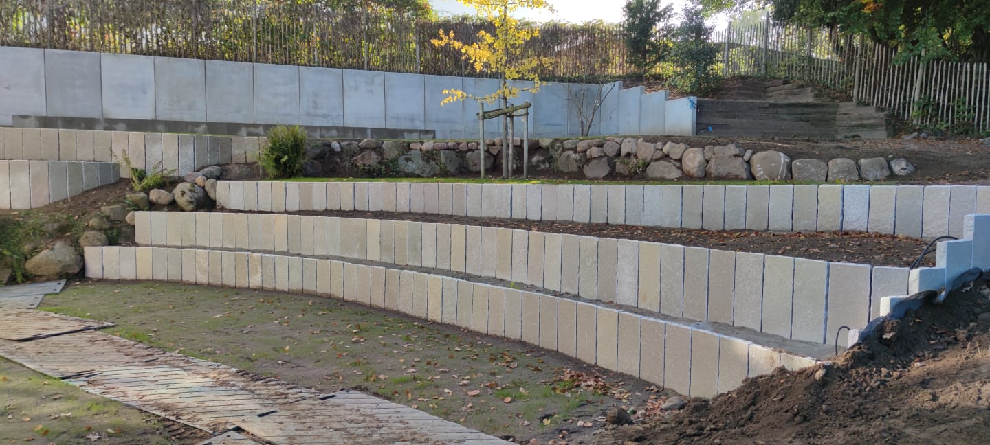 Geschwungene, abgestufte Steinstützmauern in einem mit Pflanzen, Felsen und einem jungen Baum bepflanzten Außenbereich. In unseren Referenzen finden Sie ähnliche Designs mit Hängen, Holzzäunen und üppigem Grün im Hintergrund.