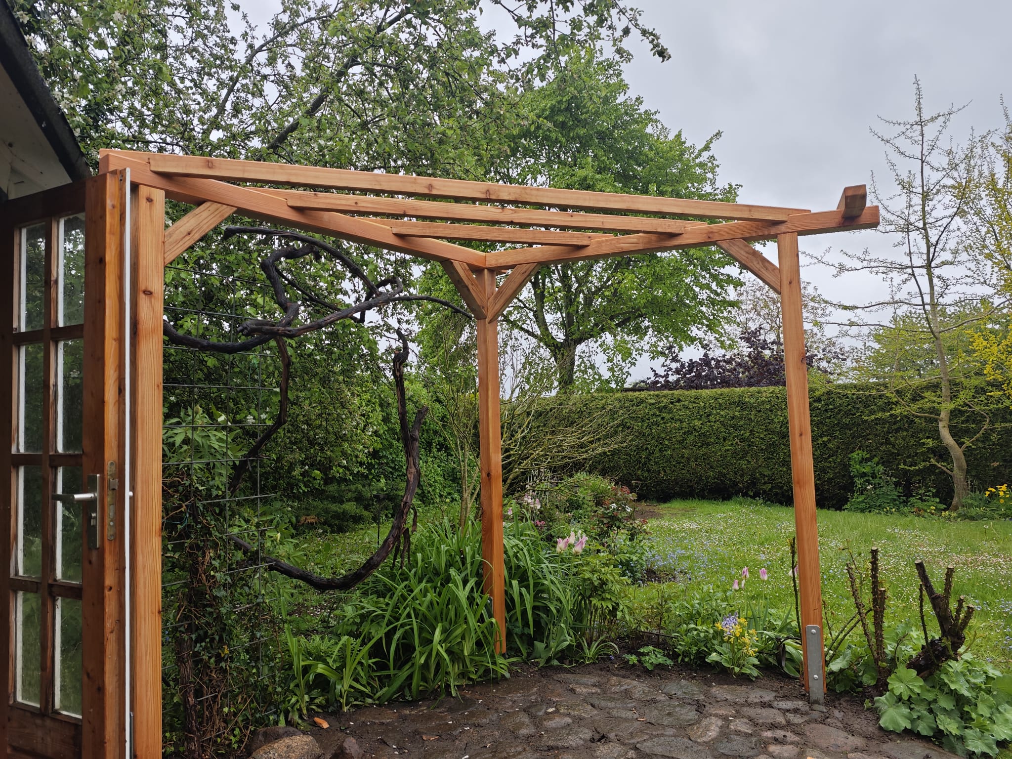 Eine hölzerne Pergola steht auf einer Steinterrasse neben einer Glastür, umgeben von üppigen grünen Pflanzen und Bäumen in einem Garten an einem bewölkten Tag.