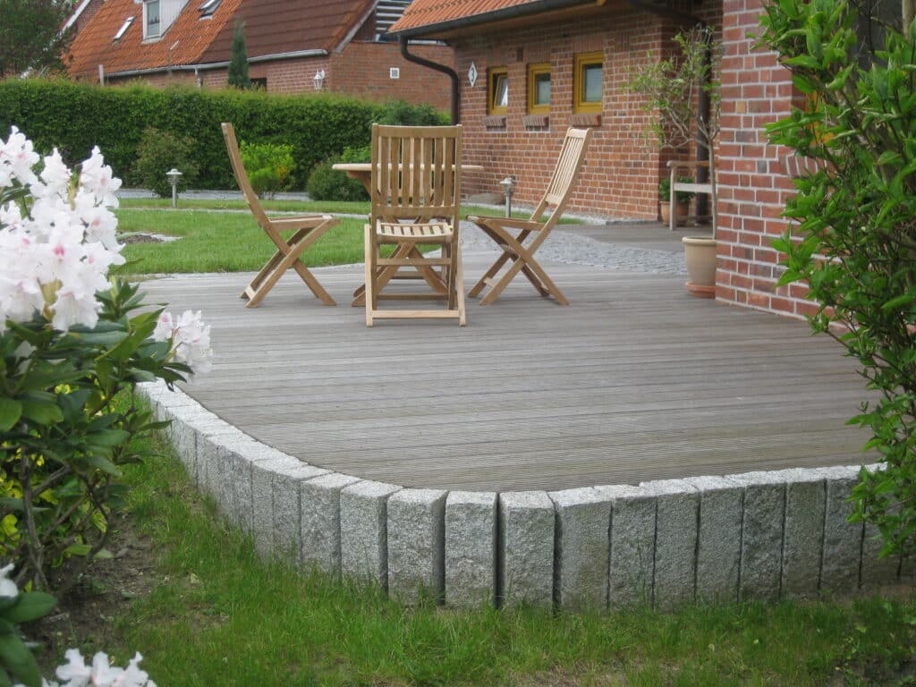 Eine Holzterrasse mit drei Holzstühlen und einem Tisch steht neben einem Backsteinhaus, umgeben von blühenden Sträuchern und grünem Gras - ein einladender Außenbereich, der zum Entspannen oder zum Sammeln von Referenzen für Ihre eigene Gartengestaltung einlädt.