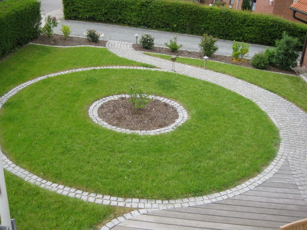Ein spiralförmiger Steinweg schlängelt sich durch einen grünen Gartenrasen und umgibt ein zentrales Blumenbeet mit kleinen Pflanzen, das von ordentlich geschnittenen Hecken und einer Straße im Hintergrund begrenzt wird.