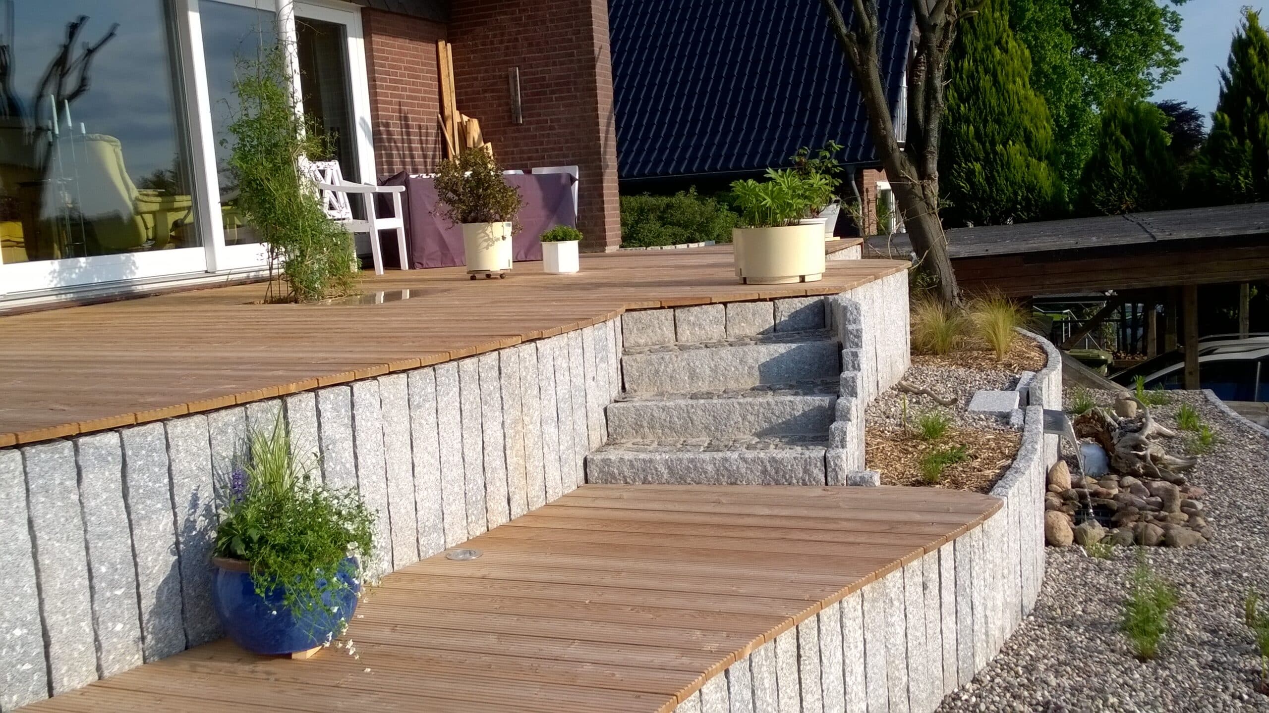 Ein moderner Innenhof mit Steinstufen, einem Holzdeck und Topfpflanzen. Der Bereich ist mit Kies, Steinen und Grünpflanzen angelegt, und ein Tisch mit Stühlen ist in der Nähe des Gebäudes zu sehen.